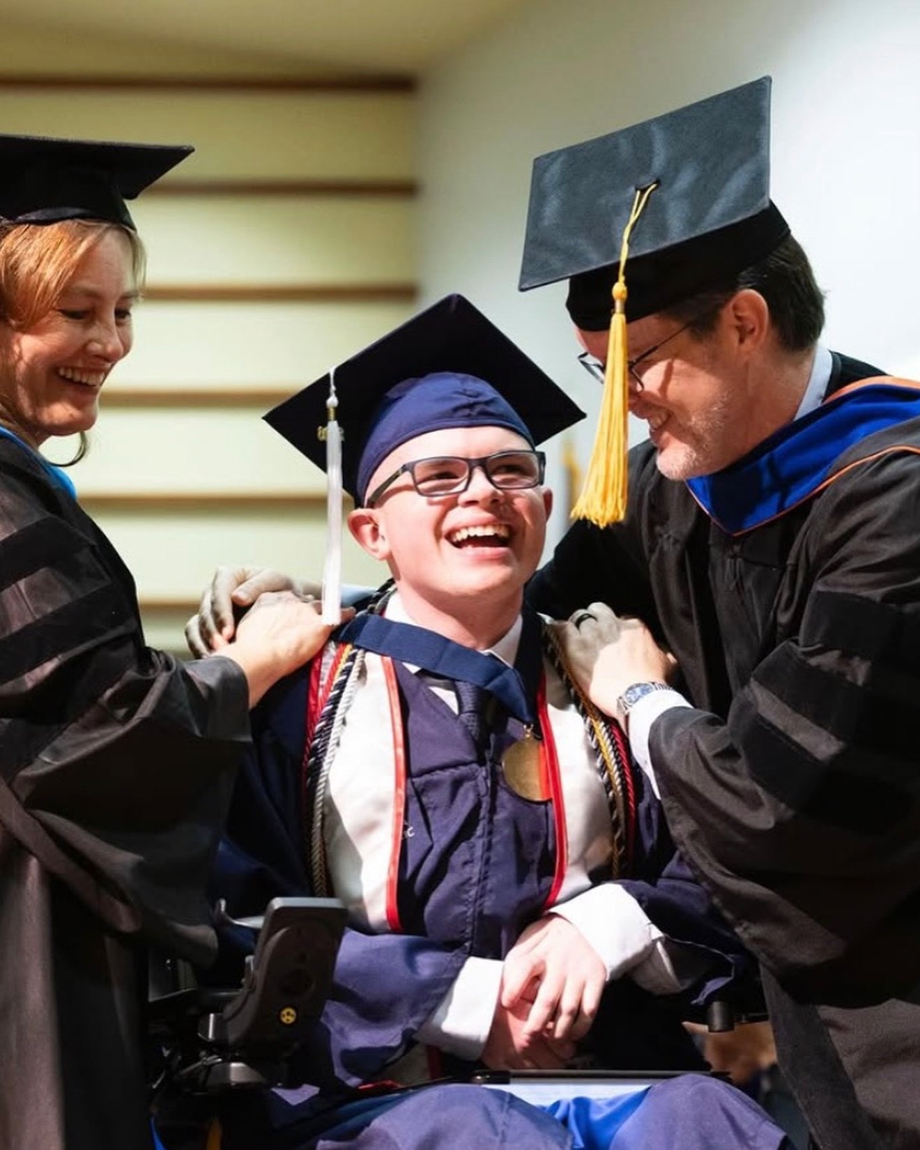 Two faculty members in academic regalia lean in to drape honor cords over a laughing Ryleigh Newman before the ceremony.