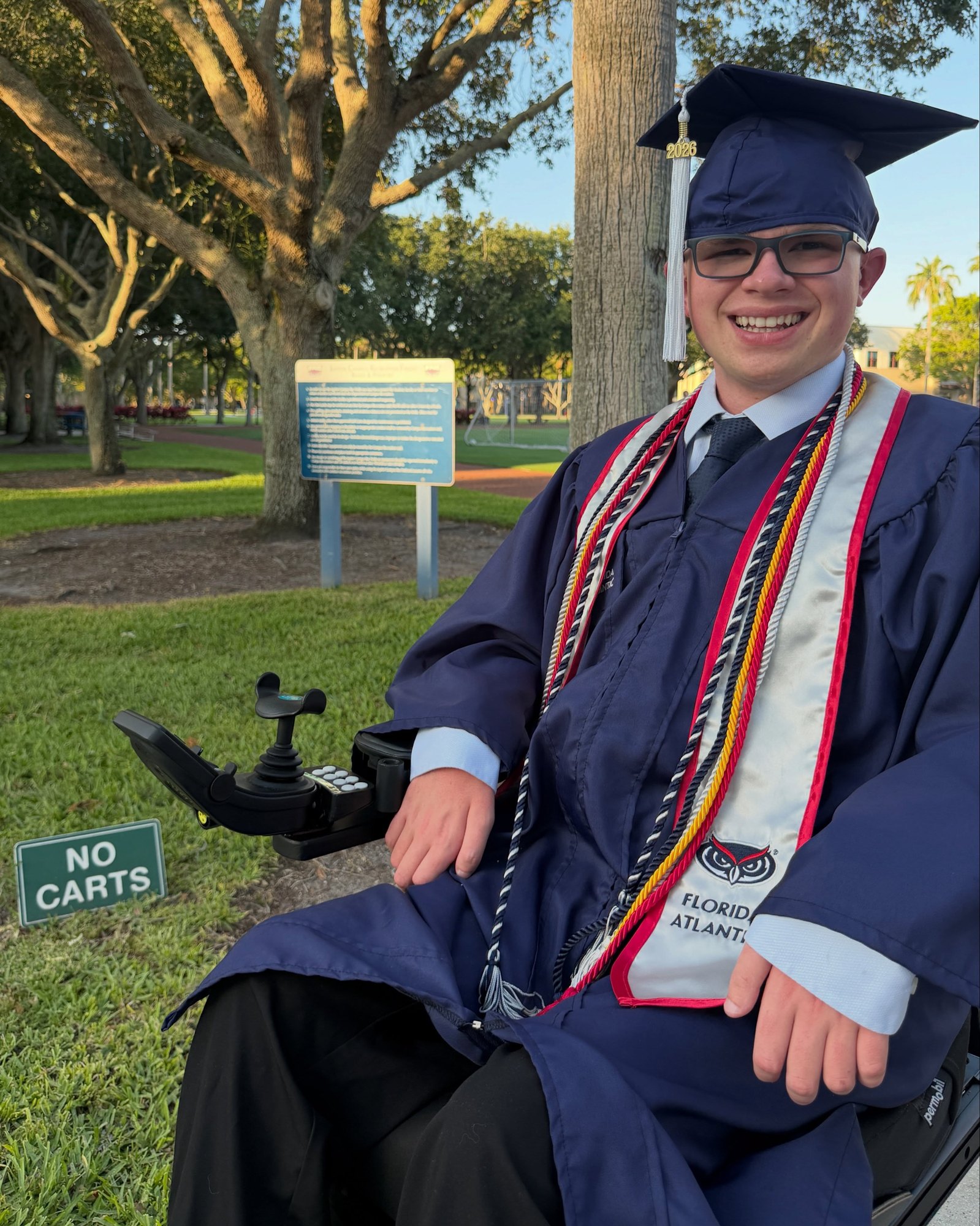 Ryleigh Newman seated in full graduation regalia under a campus tree at golden hour, honor cords and Florida Atlantic stole draped over his gown.