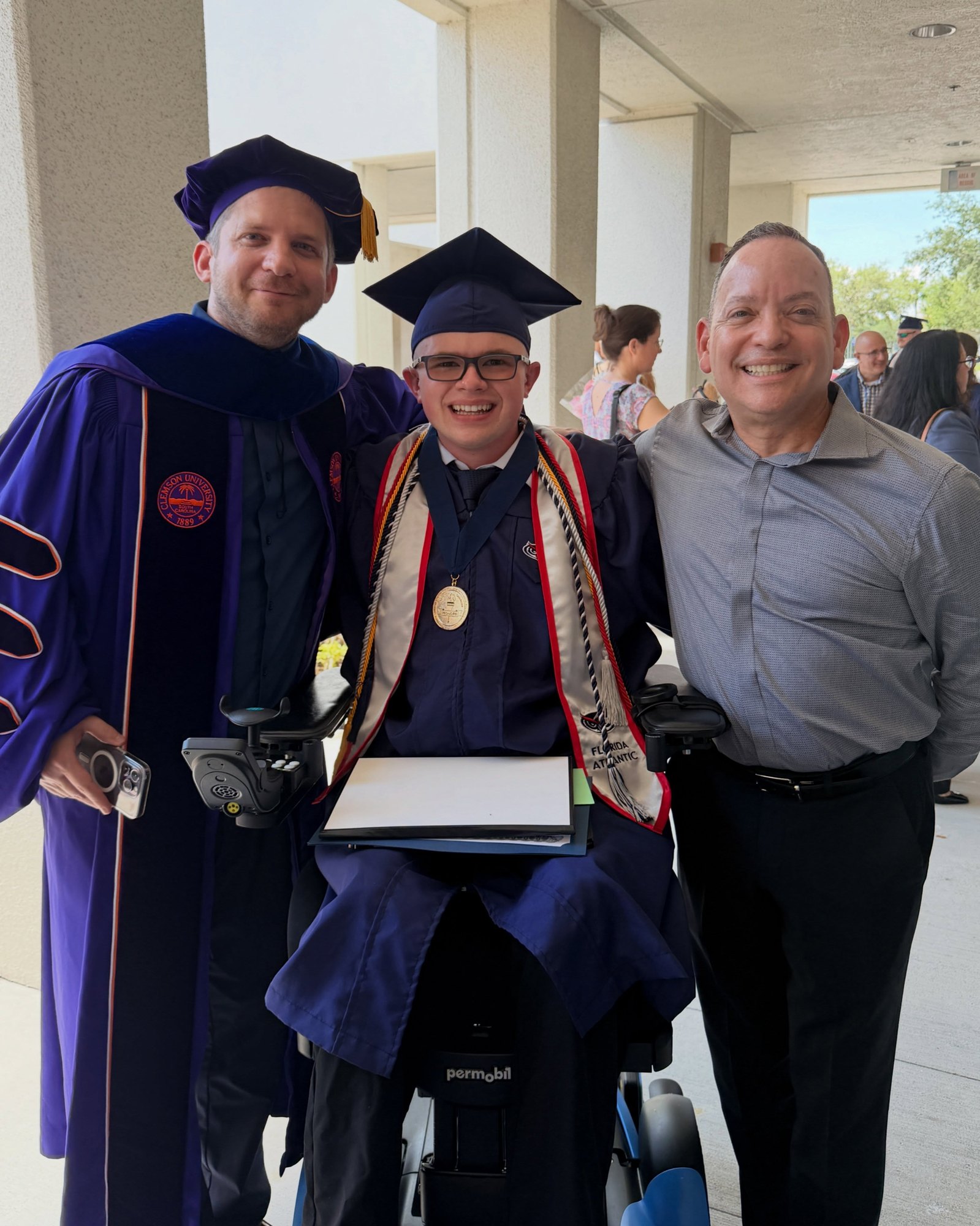 Ryleigh Newman in cap and gown holding his diploma and medallion, flanked by two faculty members in academic regalia outside the venue after the ceremony.