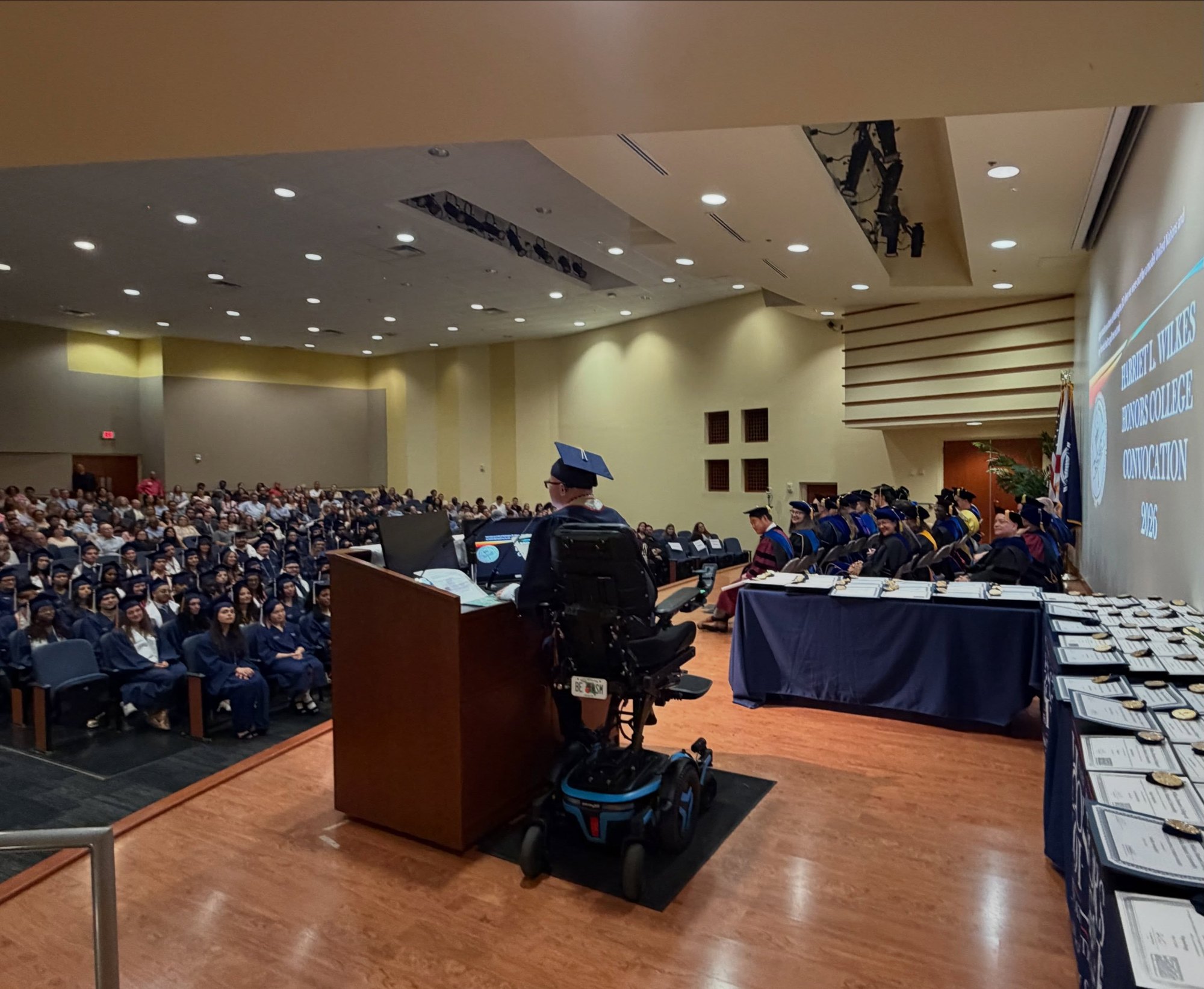 View from the stage behind Ryleigh Newman as he addresses a packed Wilkes Honors College Convocation hall, with the faculty table and rows of medallions on his right and the 2026 Convocation slide glowing on the wall.