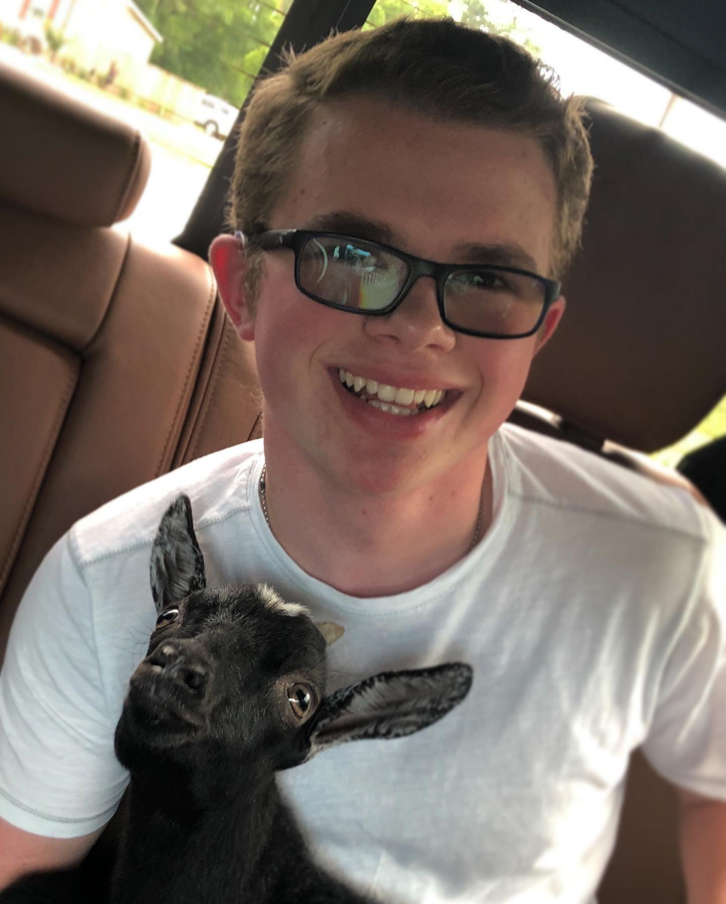 Ryleigh Newman grinning in the back seat of a car with a small black goat kid tucked against his chest, both heads tilted toward the camera.