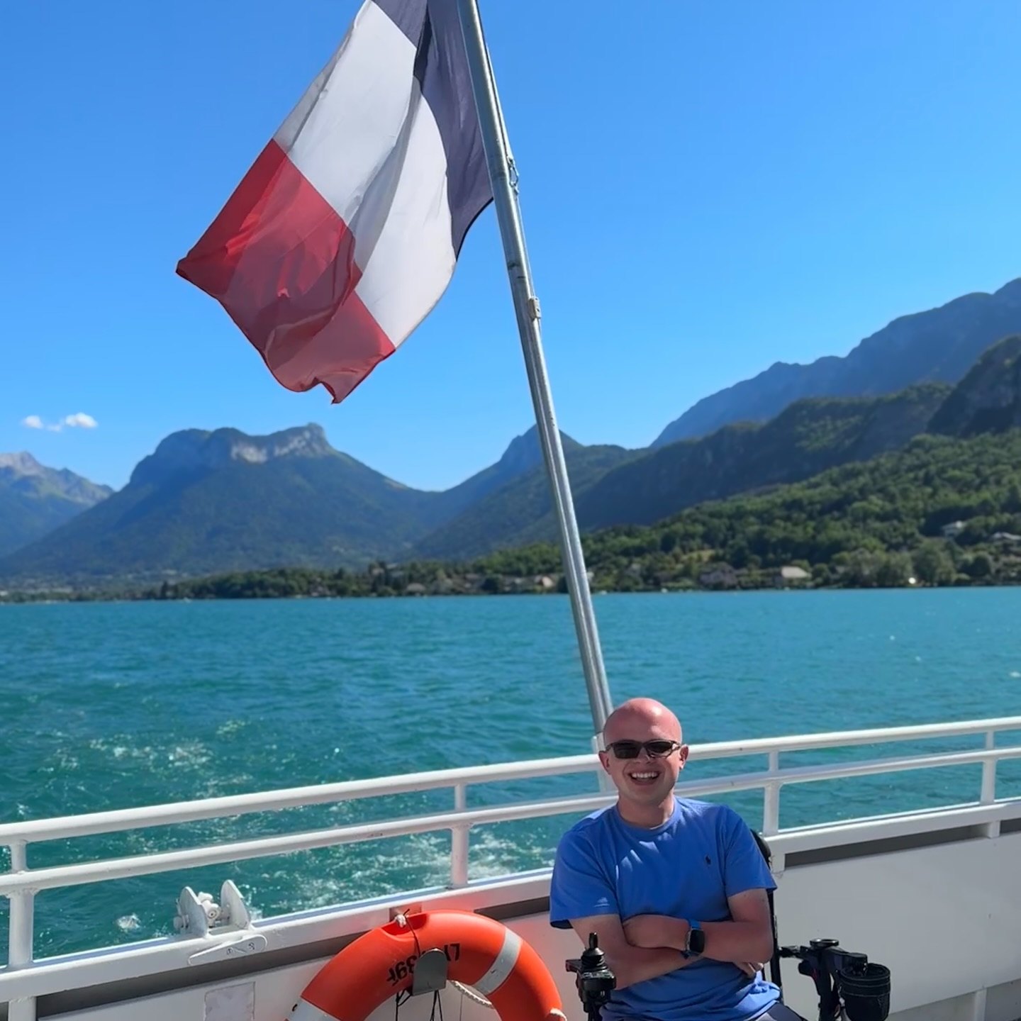 The turquoise water of Lake Annecy seen from a boat railing, with a sharply peaked green mountain rising across the lake under a clear blue sky.